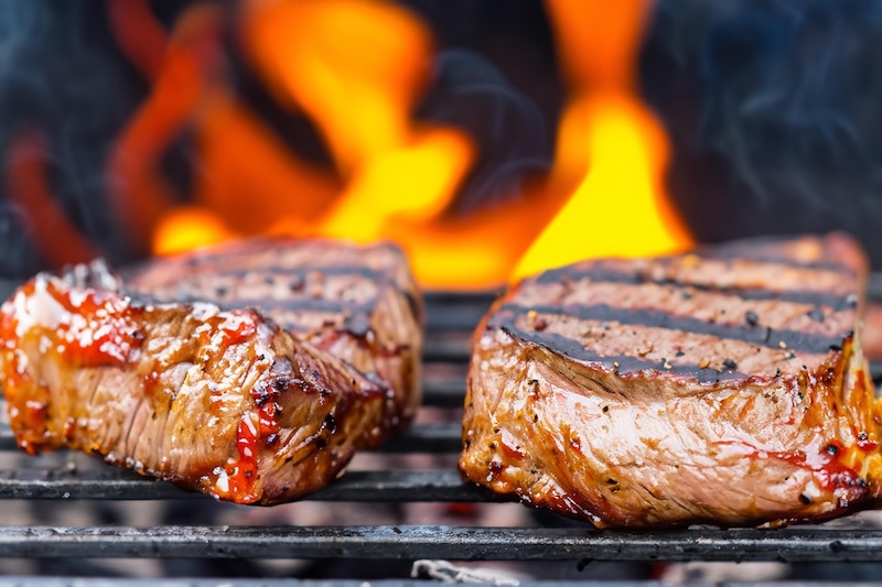 Raw steaks cooking over an open flame on a barbecue grill.
